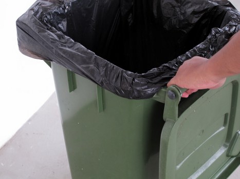 A close-up of a green plastic wheeled rubbish bin positioned on a concrete outdoor surface, with its black plastic liner lining the interior and slightly folded over the top edge. A person's hand, wearing no visible gloves, is holding the bin's handle, suggesting they are about to move or empty it. The background includes a plain white or light grey surface, indicating an outdoor or driveway setting, possibly part of a property in Southgate. This image reflects typical rubbish collection or clearance activities that Southgate House Clearance might undertake as part of their waste management services in the area. The bin appears clean with a matte finish, and the lighting is natural, highlighting the texture of the plastic and the metallic handle. Overall, the scene depicts a standard waste disposal process aligned with professional rubbish removal practices in the local vicinity.