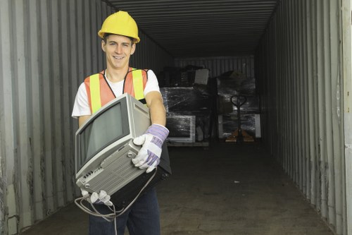 A young man wearing a yellow safety helmet, a high-visibility safety vest with orange and yellow reflective strips, a white T-shirt, and purple gloves is standing inside a corrugated metal storage container. He is smiling while holding an old, beige CRT computer monitor with a visible power cord, positioned diagonally across his body. The container's interior is dimly lit, with several large black plastic-wrapped items stacked in the background. The floor appears to be made of concrete, and the container is open at one end, revealing an outdoor environment. This scene is representative of a rubbish removal or clearance service, such as those offered by Southgate House Clearance, operating in the local area near Southgate, possibly involving the disposal of electronic waste.