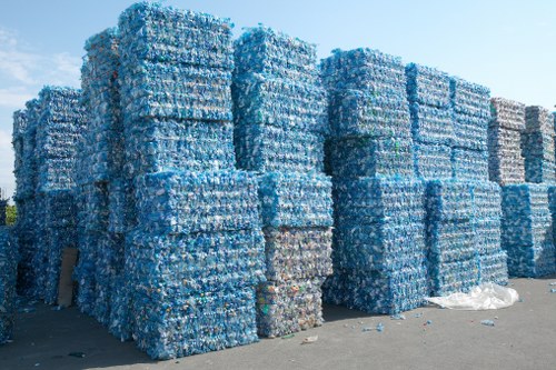 A large outdoor storage area shows towering stacks of tightly packed blue plastic bottled water bottles, arranged in uniform, rectangular blocks on a concrete ground. The bottles, with transparent and light blue shades, appear to be crushed or flattened, creating a textured, compressed appearance with some visible labels. The stacks are set against a clear, bright sky, indicating daylight, with no other objects or vehicles visible nearby. The scene reflects a waste management or recycling process, possibly associated with rubbish removal services offered by Southgate House Clearance, situated in the UK. The orderly stacks suggest preparation for recycling or disposal, highlighting the environmental management aspect of the service, with the bulk arrangement emphasizing capacity and logistical handling of plastic waste from residential or commercial sources in the local area.