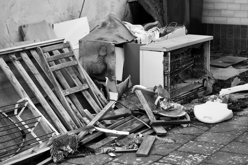 A collection of discarded household items and debris on a paved outdoor surface in front of a brick and wood-paneled wall. Visible objects include a broken wooden garden chair with slats and a weather