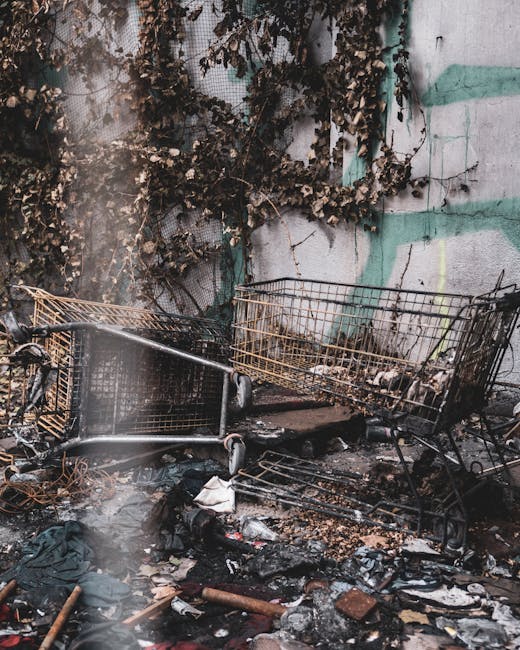 A collection of discarded household items and debris on a paved outdoor surface in front of a brick and wood-paneled wall. Visible objects include a broken wooden garden chair with slats and a weather